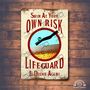 Pode incluir: Um sinal de metal de estilo vintage com um círculo vermelho e um mergulhador de desenho animado. O sinal diz "Nadar por sua conta e risco. O salva-vidas está bêbado!"