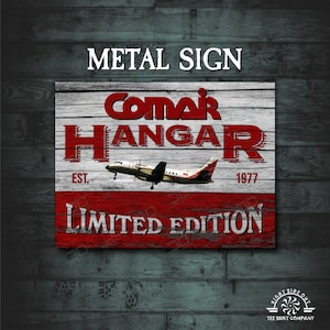 May include: Red and white metal sign with the text "ComAir Hangar Est. 1977 Limited Edition" and a small airplane in flight.