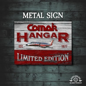 May include: A red and white metal sign with the text "Comar Hangar Est. 1977 Limited Edition". The sign features a white airplane with red accents on a woodgrain background.