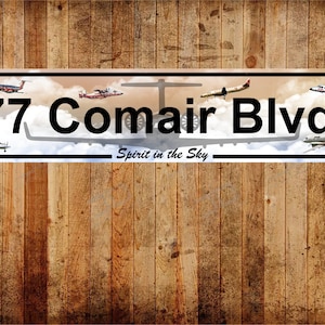 May include: A white metal sign with a blue background and a white cloud design. The sign reads "77 Comair Blvd. Spirit in the Sky". There are several small airplane silhouettes on the sign.