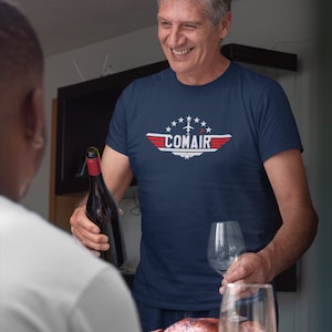 May include: A man wearing a navy blue t-shirt with a red, white, and blue airplane graphic and the text "COMAIR" printed on the front. He is holding a bottle of wine and a wine glass.