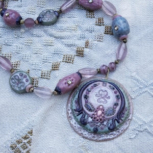 Peut inclure: Un collier vintage avec un grand pendentif rond orné d'un motif floral détaillé dans des tons de rose, lavande et bleu. La chaîne de perles comporte des perles ovales et rondes assorties, créant un accessoire délicat et orné.