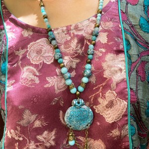 Peut inclure: Un collier turquoise et laiton avec un grand pendentif orné. Le pendentif est de forme circulaire et présente des motifs complexes. Le collier est composé de perles et est doté d'un pompon qui pend en bas.