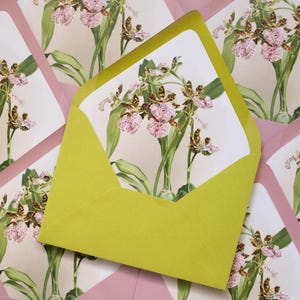 May include: A collection of stationery featuring floral illustrations. The image shows a close-up of a yellow envelope with a floral card inside, surrounded by other pink envelopes. The cards and envelopes have a botanical theme with pink and brown orchids.