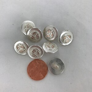 May include: Seven silver buttons with a raised design of leaves in a copper color. The buttons are approximately the size of a US penny.