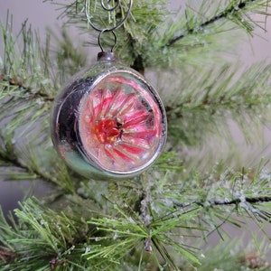 May include: A vintage silver Christmas ornament with a pink flower design inside. The ornament is hanging on a frosted artificial Christmas tree.