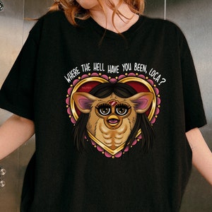 Può includere: Maglietta nera con una grafica di un cuore rosa con un contorno dorato. All'interno del cuore c'è un'immagine di un cartone animato di una creatura pelosa con occhi grandi e il testo "WHERE THE HELL HAVE YOU BEEN, LOCA?"