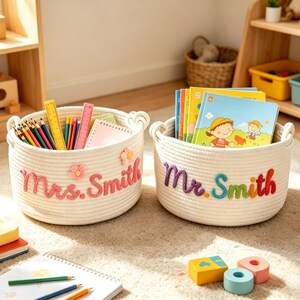Op de afbeelding: Twee crèmekleurige touwmanden met hartvormige handvatten. Eén mand heeft "Mrs. Smith" in roze geborduurde letters en bevat potloden, linialen en notitieboekjes. De andere heeft "Mr. Smith" in regenboogletters en bevat boeken.