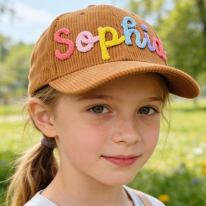 Può includere: Cappellino da baseball in velluto a coste marrone con il nome "Sophia" ricamato a lettere colorate. Il cappellino ha una visiera curva ed è indossato da una persona giovane con la pelle chiara e i capelli biondi, su uno sfondo sfocato all'aperto.