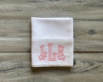 May include: White pillowcase with a pink monogram of the letters LGR. The pillowcase is folded neatly and placed on a wooden surface. The monogram is embroidered in a decorative font.