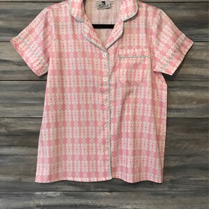 Pu&ograve; includere: Una camicia rosa con bottoni, un motivo bianco e un bordo blu chiaro. La camicia ha maniche corte e una tasca sul lato sinistro.