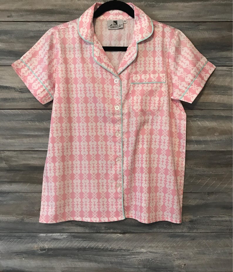 Pu&ograve; includere: Una camicia rosa con bottoni, un motivo bianco e un bordo blu chiaro. La camicia ha maniche corte e una tasca sul lato sinistro.