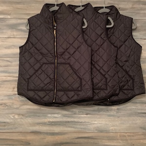 May include: Three black quilted vests with gold zippers hanging on hangers. The vests have a diamond pattern and a pocket on the front.