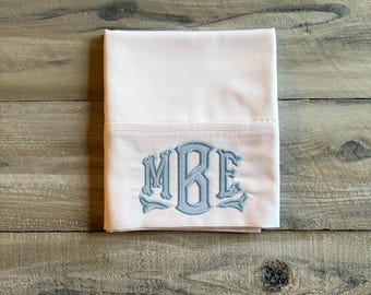 May include: White pillowcase with light blue embroidered initials MBE. The initials are in a decorative, vintage style. The pillowcase is folded and placed on a weathered, wooden surface.