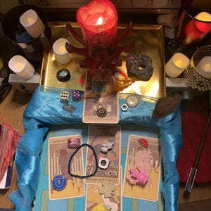 Könnte beinhalten: Eine mystische Anordnung mit Tarotkarten, Kerzen und verschiedenen dekorativen Gegenständen. Eine große rote Kerze steht auf einem goldenen Tablett, umgeben von kleineren Kerzen und anderen Objekten. Die Karten sind auf einem blauen Tuch angeordnet.