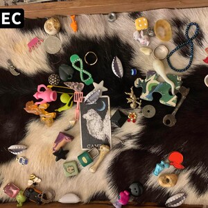 May include: A tray filled with a variety of small trinkets and toys, including a pink teapot, a green infinity symbol, a white rabbit, a yellow umbrella, a blue necklace, a white bone, a key, a puzzle piece, a tarot card with a wolf on it, and a small red and white toy. The tray is lined with a black and white cowhide.