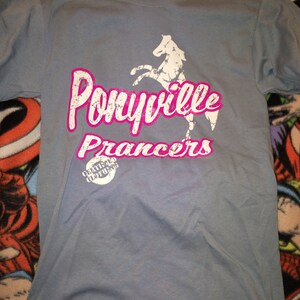 Op de afbeelding: Een lichtblauw T-shirt met een witte paard afbeelding en de tekst "Ponyville Prancers" in roze. Het shirt heeft ook een kleine witte cirkel met de tekst "Certified Clothing" in zwart.
