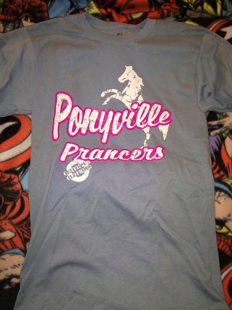 Op de afbeelding: Een lichtblauw T-shirt met een witte paard afbeelding en de tekst "Ponyville Prancers" in roze. Het shirt heeft ook een kleine witte cirkel met de tekst "Certified Clothing" in zwart.