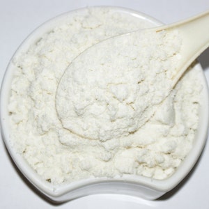 May include: A white ceramic bowl filled with a white powder, a white spoon is partially submerged in the powder.
