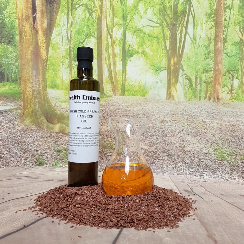 May include: A glass bottle of Health Embasso Fresh Cold Pressed Flaxseed Oil, 100% natural, with a label and a brown cap. The bottle is sitting on a pile of flax seeds in front of a blurred background of green trees.