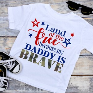 May include: White t-shirt with the text "Land of the Free because my DADDY IS BRAVE" in red, blue, and camouflage green. Includes a pair of black and white sneakers and black sunglasses.