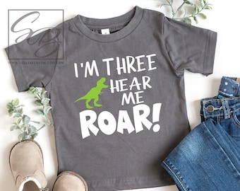 May include: A gray short-sleeved t-shirt with a green dinosaur graphic and the text "I'm Three Hear Me Roar!"