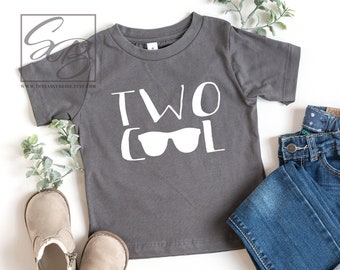 May include: A gray t-shirt with the text "TWO COOL" and sunglasses graphic, paired with blue jeans and tan boots. The outfit is styled on a white background with greenery.