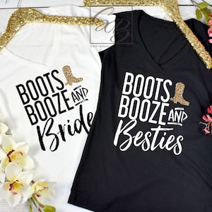 May include: Two white v-neck t-shirts with black text that reads "Boots Booze and Bride" and "Boots Booze and Besties". The shirts are hanging on gold hangers and are surrounded by white and pink flowers.