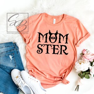 K&ouml;nnte beinhalten: Ein pfirsichfarbenes T-Shirt mit schwarzem Text, der "MOMSTER" lautet, mit einem schwarzen Auge und H&ouml;rnern &uuml;ber dem Wort "MOM".