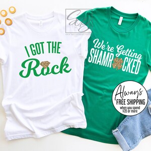 May include: Two white t-shirts with green text and a gold glitter shamrock design. The text on the first shirt says "I got the Rock" and the text on the second shirt says "We're getting Shamrocked".