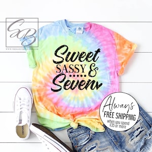 Peut inclure: Un t-shirt tie-dye avec le texte "Sweet Sassy & Seven" en noir. Le t-shirt est associ&eacute; &agrave; un short en jean et des baskets blanches. Le t-shirt pr&eacute;sente un motif en spirale dans les tons de bleu, jaune, orange et rose.