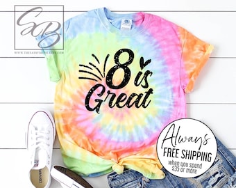 May include: A tie-dye t-shirt with the text "8 is Great" in black, paired with denim shorts and white sneakers. The shirt features a spiral pattern in shades of blue, green, yellow, orange, and pink.