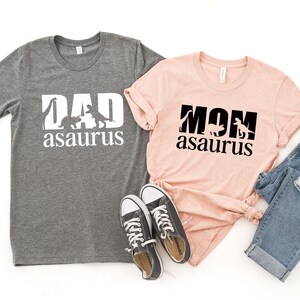 May include: Two t-shirts with a dinosaur graphic and the text "DAD asaurus" and "MOM asaurus". The shirts are grey and pink and are laid out on a white background.