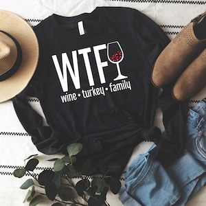 K&ouml;nnte beinhalten: Schwarzes Langarmshirt mit dem wei&szlig;en Text "WTF wine &bull; turkey &bull; family" und einer Weinglas-Grafik. Ein Strohhut, braune Stiefel und blaue Jeans sind im Hintergrund.