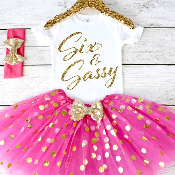 May include: A white short-sleeved bodysuit with gold glitter text reading Six & Sassy, paired with a bright pink tulle tutu skirt featuring gold polka dots and a gold sequin bow, plus a matching pink headband with a gold sequin bow.