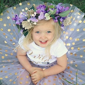 Puede incluir: Una ni&ntilde;a peque&ntilde;a con una camisa blanca con el texto "Wild One" y una flecha, una falda de tul lavanda con lunares dorados y una corona de flores con flores moradas y blancas.