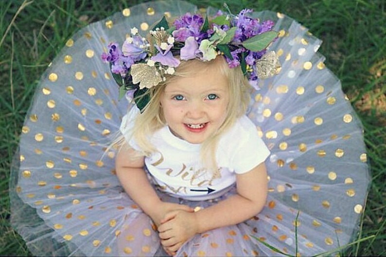 Puede incluir: Una ni&ntilde;a peque&ntilde;a con una camisa blanca con el texto "Wild One" y una flecha, una falda de tul lavanda con lunares dorados y una corona de flores con flores moradas y blancas.