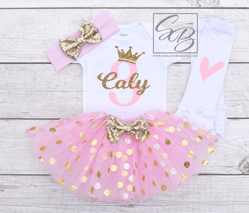 Pu&ograve; includere: Un completo per il compleanno di una bambina. Il set include un body bianco con una corona glitterata dorata e il nome "Caly" in glitter dorati, un tut&ugrave; rosa con pois dorati, una fascia rosa con un fiocco dorato e leggings bianchi con un cuore rosa.