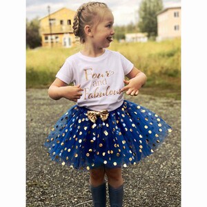 May include: A young girl wearing a white t-shirt with gold glitter text that says "Four and Fabulous" and a blue tulle skirt with gold polka dots.
