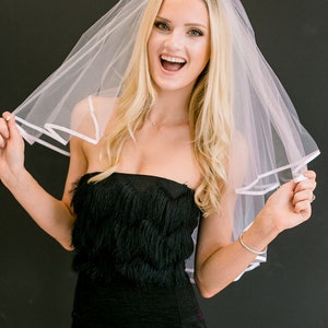 May include: A woman wearing a white bridal veil with a black strapless dress. She is smiling and holding the veil up.