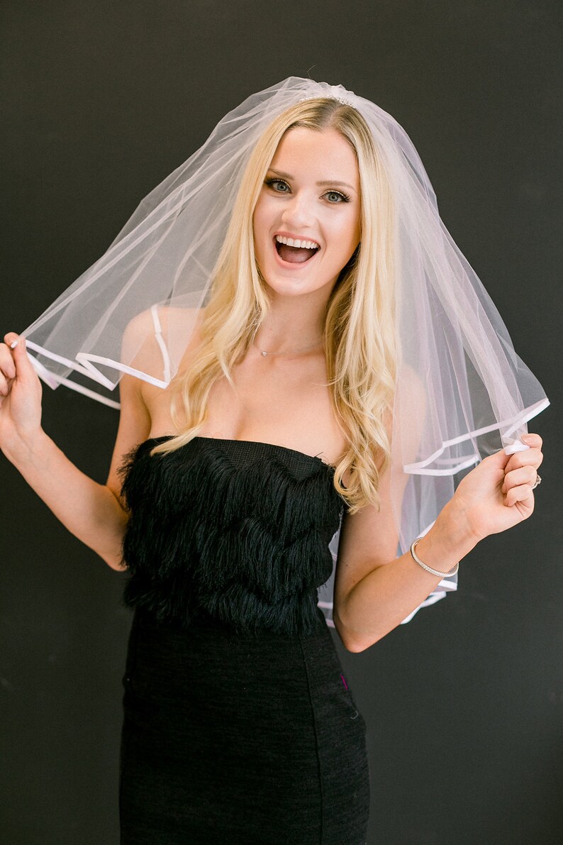 May include: A woman wearing a white bridal veil with a black strapless dress. She is smiling and holding the veil up.