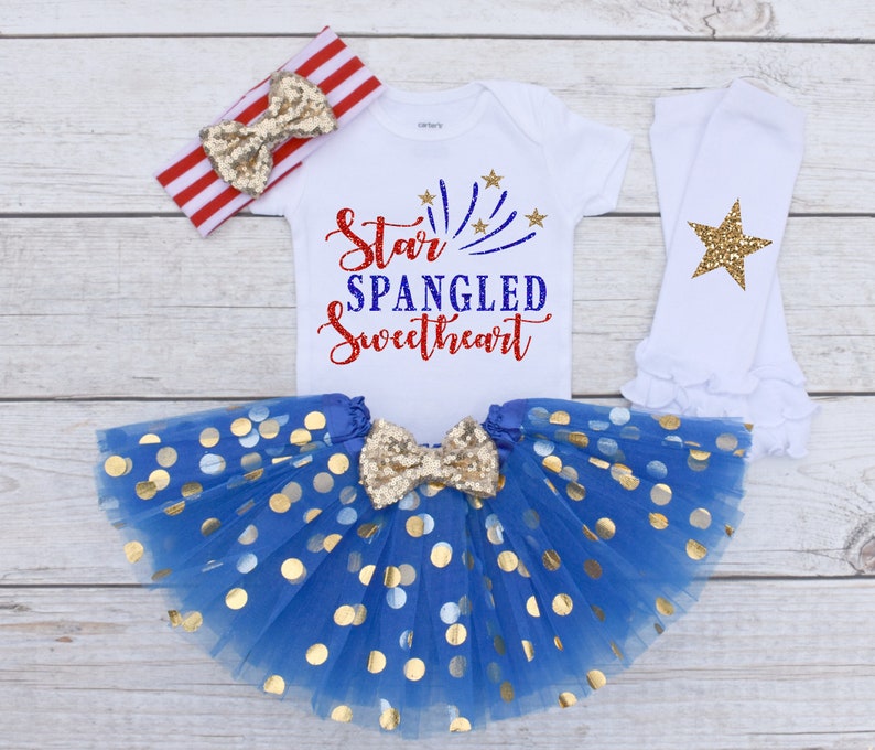 May include: A baby girl's 4th of July outfit with a white bodysuit that says "Star Spangled Sweetheart" in red, blue, and gold glitter. The outfit also includes a blue tulle skirt with gold polka dots, a red and white striped headband with a gold glitter bow, and white leg warmers with a gold glitter star.