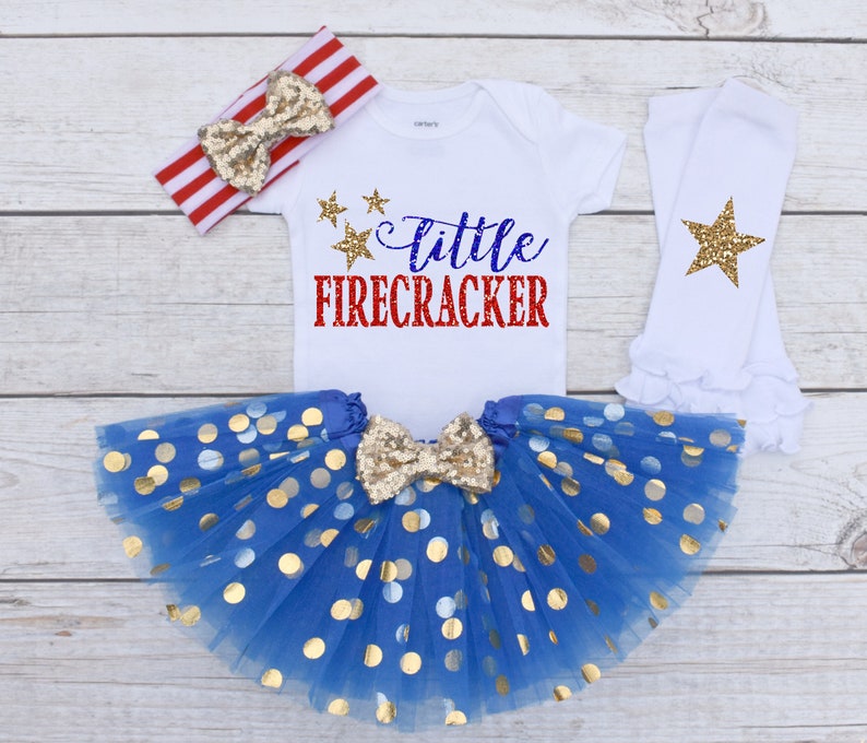 May include: A patriotic baby outfit set featuring a white onesie with "Little Firecracker" text, a blue tutu skirt with gold polka dots, a red and white striped headband, and white leg warmers with gold stars.