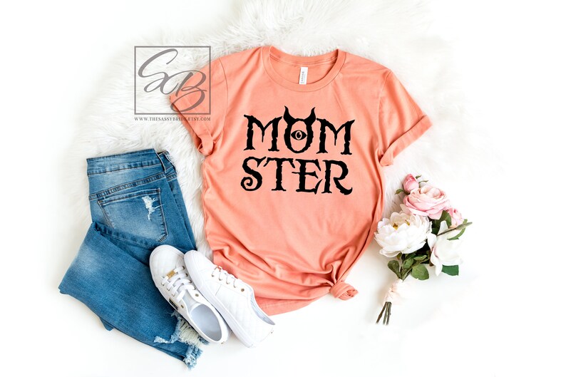 K&ouml;nnte beinhalten: Ein pfirsichfarbenes T-Shirt mit schwarzem Text, der "MOMSTER" lautet, mit einem schwarzen Auge und H&ouml;rnern &uuml;ber dem Wort "MOM".