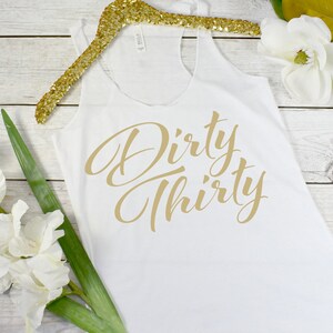May include: White tank top with the text "Dirty Thirty" in gold lettering.