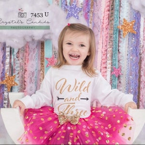 Puede incluir: Una ni&ntilde;a peque&ntilde;a con una camiseta blanca de manga larga con letras doradas brillantes que dicen "Wild and Three" y una falda de tul rosa con lunares dorados. Est&aacute; sonriendo y mirando a la c&aacute;mara.