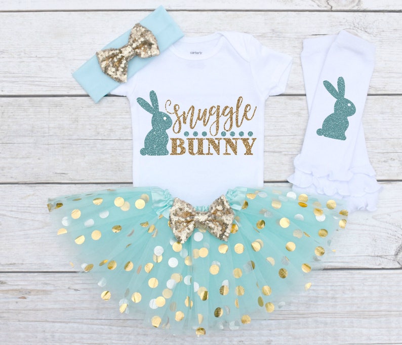 May include: A baby girl's Easter outfit with a white bodysuit that says "Snuggle Bunny" in gold glitter, a mint green tulle skirt with gold polka dots, a mint green headband with a gold glitter bow, and white leg warmers with a teal glitter bunny on each.