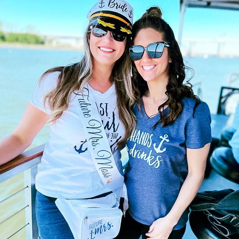 May include: Two women wearing white and blue shirts with nautical themed text and an anchor graphic. One woman is wearing a white sash with the text "Future Mrs. (last name)" and a captain's hat with the text "Bride".