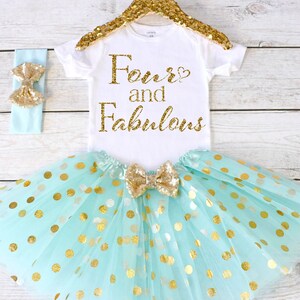 May include: A baby girl's outfit for a fourth birthday. The outfit includes a white bodysuit with gold glitter text that says "Four and Fabulous", a mint green tulle skirt with gold polka dots, and a mint green headband with a gold glitter bow.