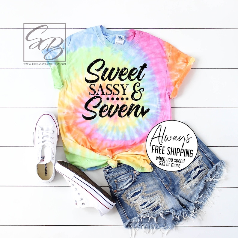 Peut inclure: Un t-shirt tie-dye avec le texte "Sweet Sassy & Seven" en noir. Le t-shirt est associ&eacute; &agrave; un short en jean et des baskets blanches. Le t-shirt pr&eacute;sente un motif en spirale dans les tons de bleu, jaune, orange et rose.